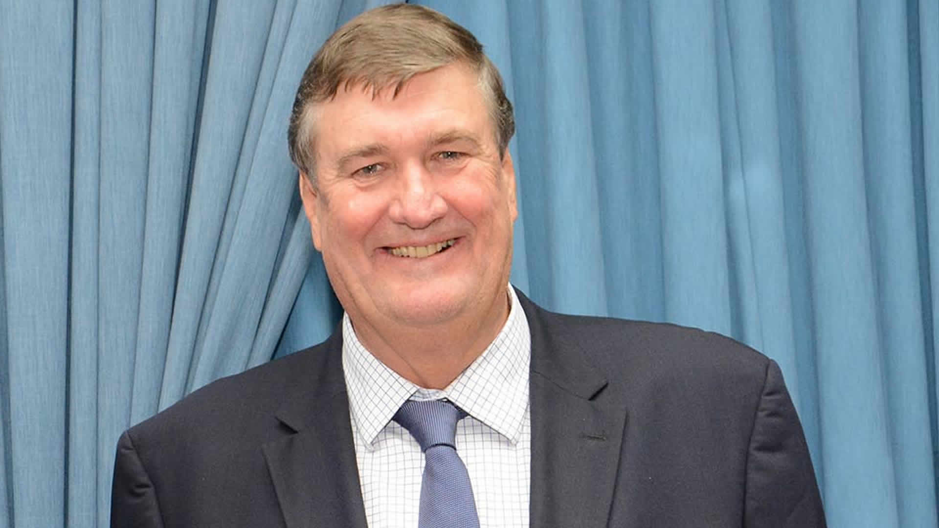 Arnaud Dalais smiling in a dark suit and blue tie in front of a blue curtain at an official function