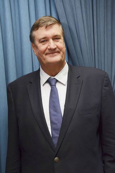 Full-length standing portrait of Arnaud Dalais in a dark suit in front of a blue curtain